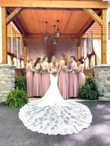 Brides dress and train with bridesmaids in front of pavilion.