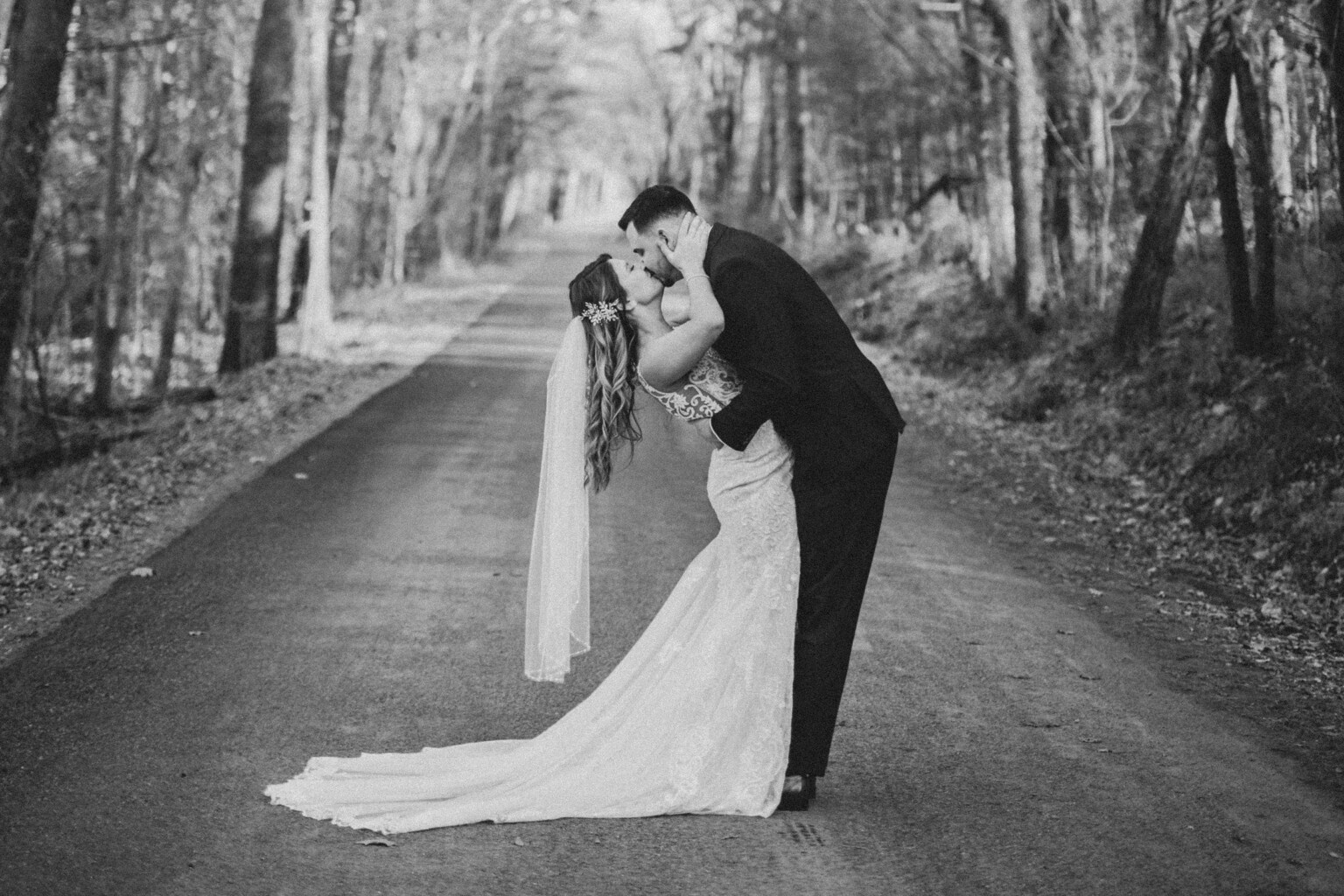 Black and white photo of couple kissing on the tree lined road